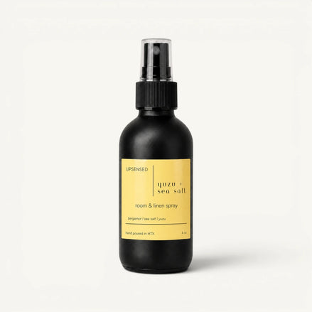 Upsensed Yuzu + Sea Salt room and linen spray in a matte black 4 oz bottle with a pale yellow label, bergamot, sea salt, and yuzu scent notes, on a plain white background.
