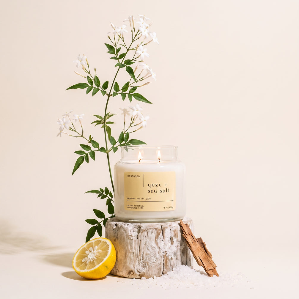 Upsensed Yuzu + Sea Salt candle burning on a weathered wood stump, styled with jasmine branches, a halved lemon, driftwood, and scattered sea salt.
