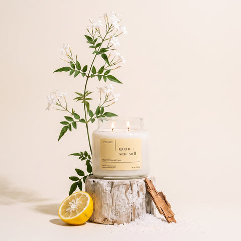 Upsensed Yuzu + Sea Salt candle burning on a weathered wood stump, styled with jasmine branches, a halved lemon, driftwood, and scattered sea salt.