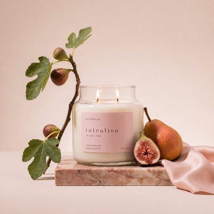 Upsensed Intention candle burning on a pink marble slab, styled with a fig branch, a halved fig, a pear, and draped pink silk fabric.