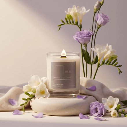 Upsensed Veil 11oz scented candle burning with wood wick surrounded by white freesia and violet flowers on soft cashmere fabric - coconut apricot wax candle hand poured in Houston