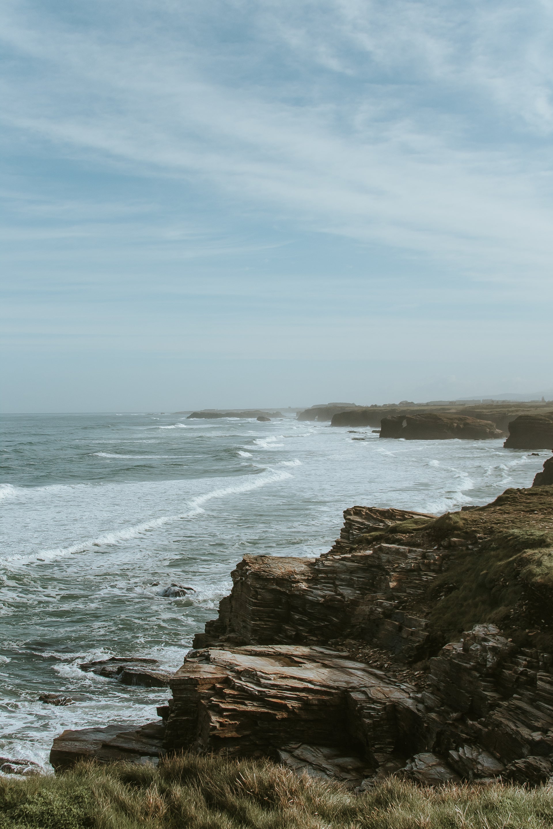 Rugged coastal cliffside with crashing ocean waves under an overcast sky — editorial background image for Upsensed Undertow candle, a marine mineral and driftwood scented candle hand-poured in Houston